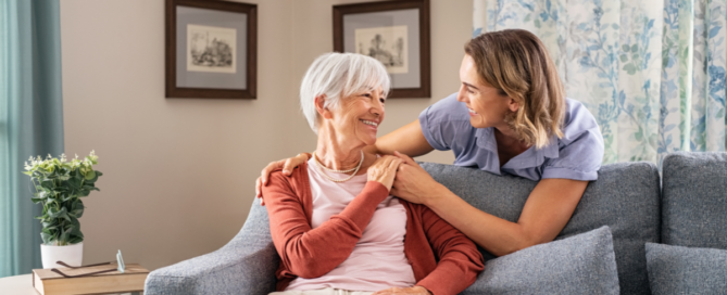 Smiling caregiver with hand over the shoulder of a happy elderly woman sitting on the couch, representing the results of quality home care services.
