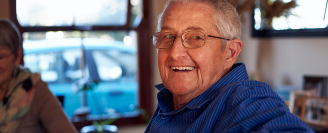 A happy, smiling senior man is sitting at a table and looking over his shoulder, enjoying aging in place as a result of successful of long-distance caregiving.