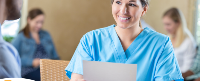 A woman in scrubs holds a paper while interviewing with someone else, representing how a solid caregiver resume can get you to an interview.